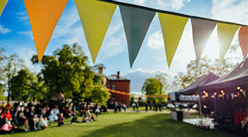 A lively outdoor festival scene with colorful bunting flags strung across a sunlit park, where people gather and enjoy a communal celebration under clear skies